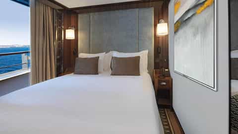 A bed with a balcony view of the ocean and coastline - World Traveller, Atlas Ocean Voyages