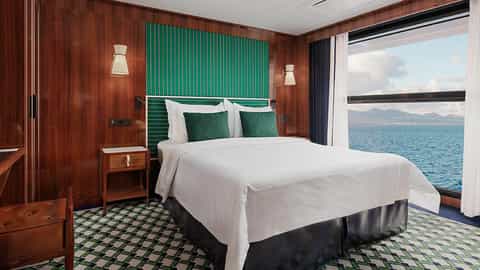 A well-appointed suite bedroom with a large window overlooking the ocean - World Traveller, Atlas Ocean Voyages