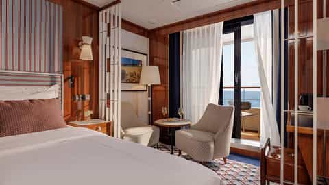 A suite with a bed, chairs, and a balcony overlooking the ocean - World Traveller, Atlas Ocean Voyages