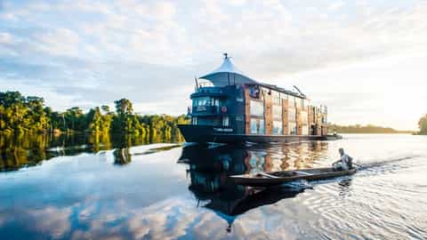 A riverboat sails on a calm river with a canoe nearby - Aria Amazon, Ponant Cruises