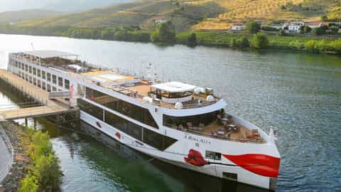 A river ship is docked on a calm river with hills in the background - Arosa Alva, A Rosa Cruises