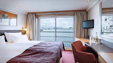 A bed and desk in a suite with a large window - Arosa Clea, A Rosa Cruises