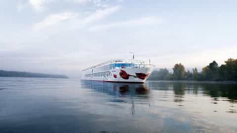 A river ship sails on calm water near a forested shore - Arosa Mia, A Rosa Cruises