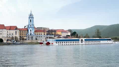 A white river ship sails past a European town with a blue-domed church - Arosa Mia, A Rosa Cruises