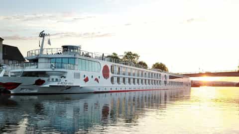 A river ship is docked near a building at sunset - Arosa Viva, A Rosa Cruises