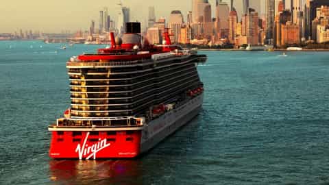A large cruise ship sails past a city skyline - Brilliant Lady, Virgin Voyages