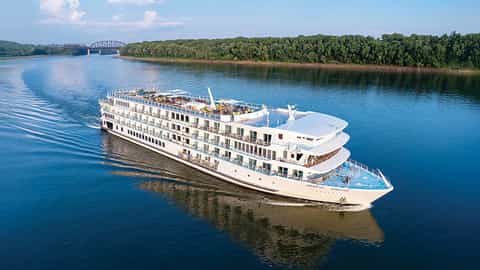 A river cruise ship sails on a wide blue river - ACL American Serenade, American Cruise Lines
