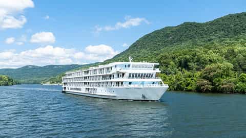 A white river ship cruises on a blue river with green hills - ACL American Symphony, American Cruise Lines