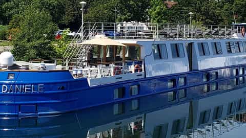 A blue and white river boat is docked on calm water - Daniele barge, CroisiEurope