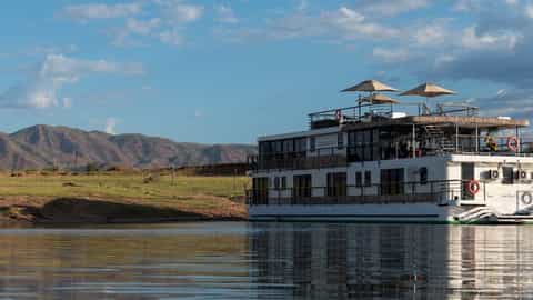 A river ship is docked by a grassy shore with mountains - African Dream, CroisiEurope