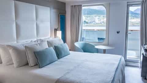 A well-appointed suite bedroom with a view of a coastal town - Le Boreal, Ponant Cruises