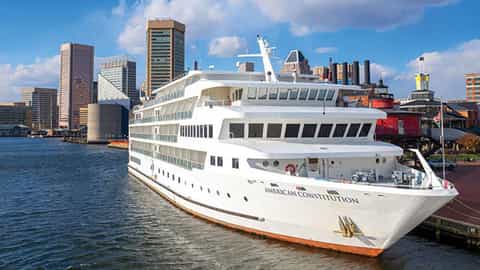 A white cruise ship is docked in a harbor with a city skyline - ACL American Constitution, American Cruise Lines