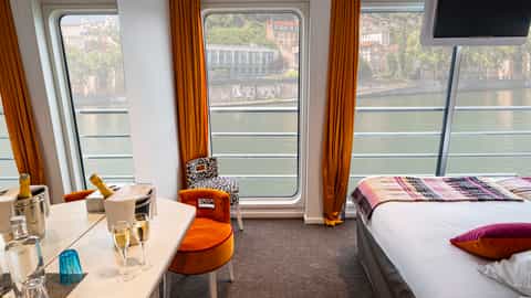 A suite with a bed, seating, and a river view - ms Camargue, CroisiEurope