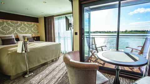 A suite with a bed, seating area, and balcony overlooking a river - MS Thomas Hardy, Riviera Travel