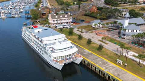 A large white riverboat is docked at a pier next to a town - ACL American Glory, American Cruise Lines