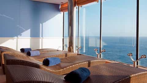 Wicker lounge chairs with blue towels face the ocean through large windows - MSC Lirica, MSC Cruises
