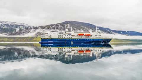 A ship sails on calm water with misty mountains behind - National Geographic Explorer, Lindblad Expeditions