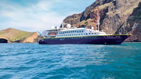 A ship sails near a rocky, vegetated coastline - National Geographic Islander 2, Lindblad Expeditions