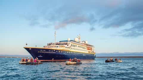 A large ship is anchored offshore with zodiacs nearby - National Geographic Islander 2, Lindblad Expeditions