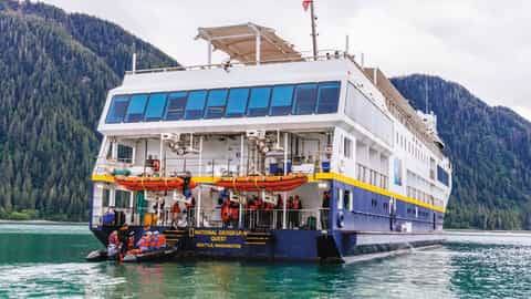 A ship sails on the water near a forested mountain - National Geographic Quest, Lindblad Expeditions