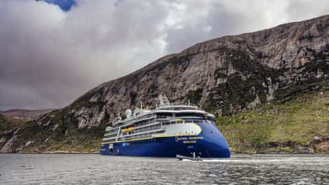 A large blue ship sails near a rocky, green mountain - National Geographic Resolution, Lindblad Expeditions