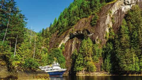 A ship sails through a narrow waterway surrounded by forested cliffs - National Geographic Sea Bird, Lindblad Expeditions