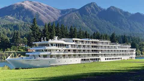 A white river ship is docked near a grassy shore - ACL American Harmony, American Cruise Lines